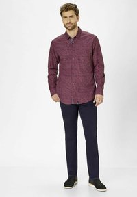 Langharige bordeauxrode shirt met een geometrisch patroon, met knoopsluiting bij de kraag, en gecombineerd met donkerblauwe broek en zwarte instap schoenen.