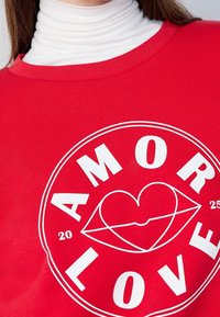 Primer plano de una persona que lleva un suéter rojo con un diseño circular blanco que muestra "AMOR LOVE" y un gráfico de labios en forma de corazón.