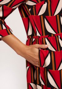 Czerwona sukienka z geometrycznym wzorem w czarne, białe i brązowe akcenty. Widać krótkie rękawy i boczne kieszenie. Gładka struktura tkaniny.