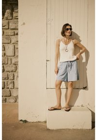 White knitted tank top, light blue shorts, black flip-flops, and sunglasses; standing against a textured stone wall and pale door.