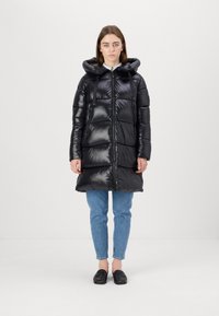 Zwarte gewatteerde jas met een glanzende afwerking, voorzien van een capuchon en een halve rits, gecombineerd met blauwe jeans en zwarte schoenen.
