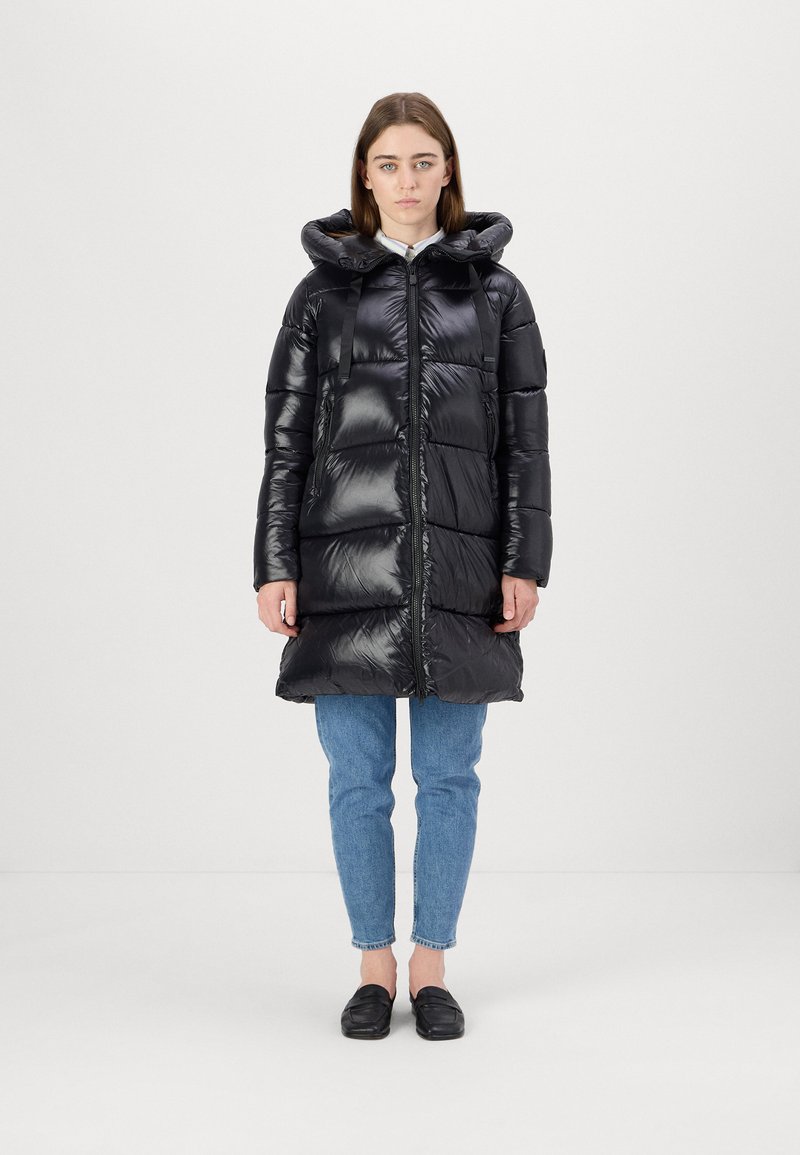 Zwarte gewatteerde jas met een glanzende afwerking, voorzien van een capuchon en een halve rits, gecombineerd met blauwe jeans en zwarte schoenen.