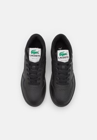 Par crnih Lacoste tenisica s vezicama, bijele oznake s logom koji prikazuje zeleni krokodil i tekst "Lacoste od 1933.", gledano odozgo.