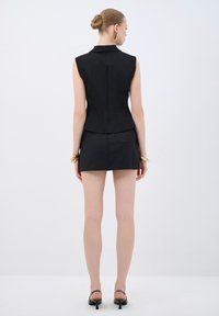 Schwarze ärmellose maßgeschneiderte Weste mit tailliertem Schnitt, kombiniert mit einem kurzen Rock. Glatte Textur, minimales Design, mit einer Gesäßtasche.
