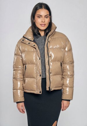 Vrouw draagt een glanzende beige donsjas over een grijze trui en een zwarte rok met een voorste split, staand tegen een effen lichte achtergrond.