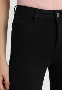 Vaqueros de mezclilla negra con un corte ajustado, que cuentan con bolsillos delanteros, un cierre de botón y detalles de costura visibles a lo largo de las costuras.