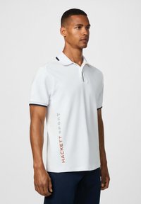 Muž v bielej polokošeli s krátkym rukávom a čiernym lemovaním, na ľavej strane s vertikálne potlačeným nápisom "Hackett Sport", v kombinácii s tmavými nohavicami.