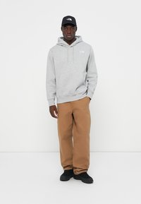 Sweatshirt gri cu glugă, dotat cu un buzunar frontal și un logo, asortat cu pantaloni bej și adidași negri. Modelul poartă o șapcă neagră. Design simplu, casual.