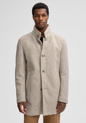 Cappotto beige a spina di pesce con colletto alto, chiusura con bottoni e tasche laterali, realizzato in un tessuto misto lana texturizzato. Vestibilità sartoriale.