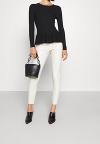 Black ribbed long-sleeve top with peplum hem, paired with white fitted pants and pointed black mules. Holding a small black handbag with chain handle.