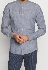 Man wearing a buttoned, long-sleeve, light blue collarless shirt and navy blue pinstripe pants, clasping hands at waist.