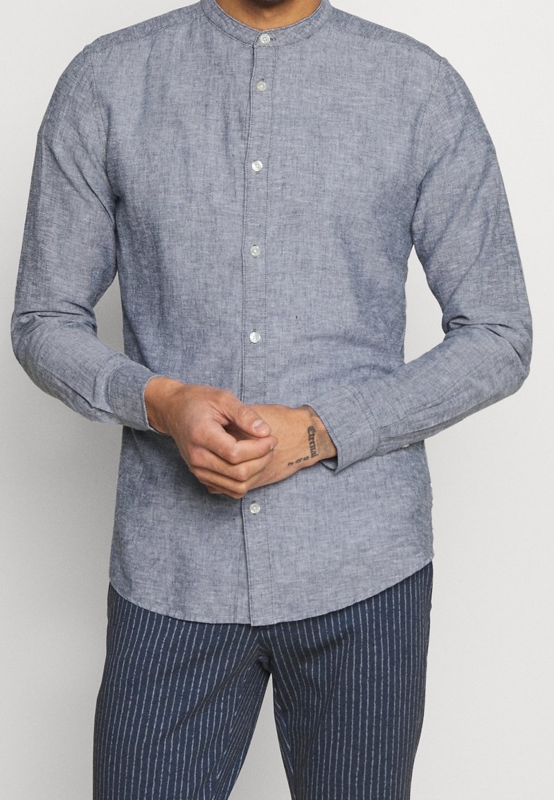 Man wearing a buttoned, long-sleeve, light blue collarless shirt and navy blue pinstripe pants, clasping hands at waist.