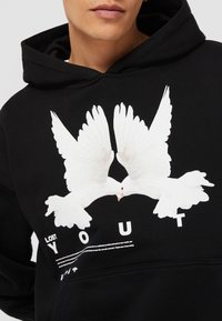 Black hoodie with a prominent white dove graphic, featuring large wings. Text "LOST YOU" and additional details printed below the graphic.