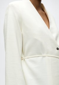 Λευκό σακάκι με υφή, βαθύ V-λαιμόκοψη και ένα μαύρο κουμπί, φορεμένο από άτομο, κομμένο στο ύψος του στήθους και των ώμων.