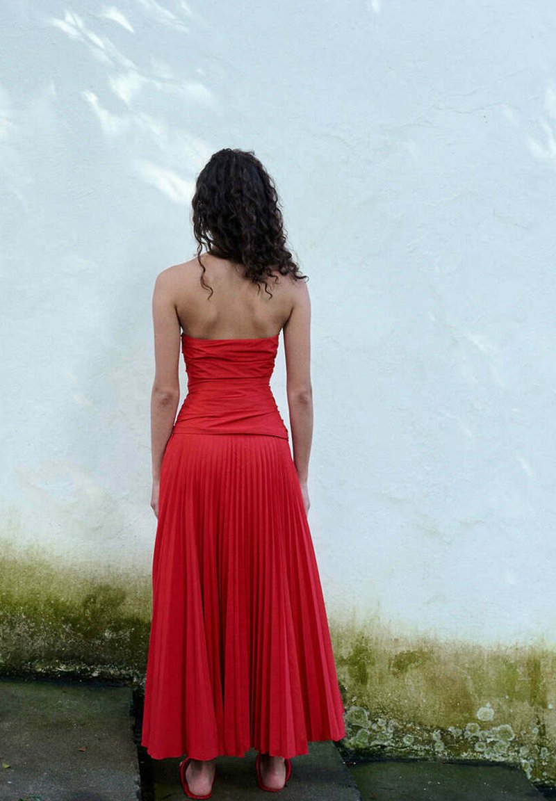 Femme aux cheveux bouclés se tient face à un mur blanc, portant une robe rouge plissée sans bretelles et des chaussures rouges sur un sol mouillé.