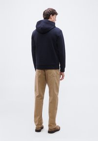 Άνδρας με την πλάτη γυρισμένη, φορούν ένα μπλε σκούρο hoodie, μπεζ παντελόνι και καφέ παπούτσια, στέκεται μπροστά σε ένα απλό ανοιχτό γκρι φόντο.