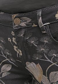 Primer plano de jeans de mezclilla negra con estampado floral beige y un botón de metal grabado con "Guess 1981."