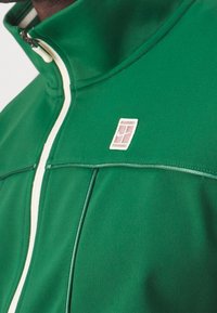 Groene ritsjack met crèmekleurige accenten en een gestructureerde logo patch. Heeft een hoge kraag en een slanke pasvorm, die de gladde stofstructuur toont.