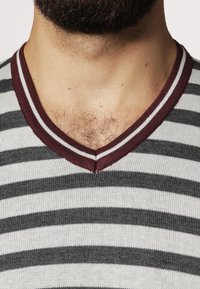 Grå och vit randig V-ringad tröja med mörkröda accenter. Har horisontella ränder och en mjuk, stickad textur.