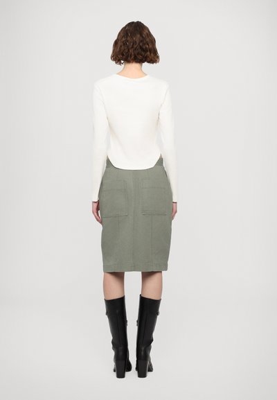 White long-sleeve top paired with a green knee-length skirt featuring two back pockets and black knee-high boots. Smooth fabric with a structured design.