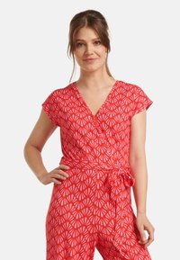 Rode jumpsuit met korte mouwen, wikkelontwerp en een gestrikt middel. Heeft een roze schelpprint op lichte stof.