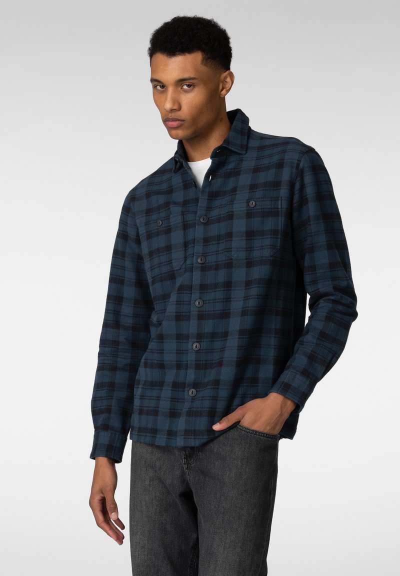 Blå og svart rutete flanellskjorte med lange ermer, knappeclosure og to brystlommer, brukt over en hvit skjorte.