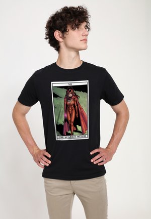 Jeune homme aux cheveux bouclés portant un t-shirt noir avec une illustration de Scarlet Witch en costume rouge et cape de style bande dessinée, debout les mains sur les hanches.