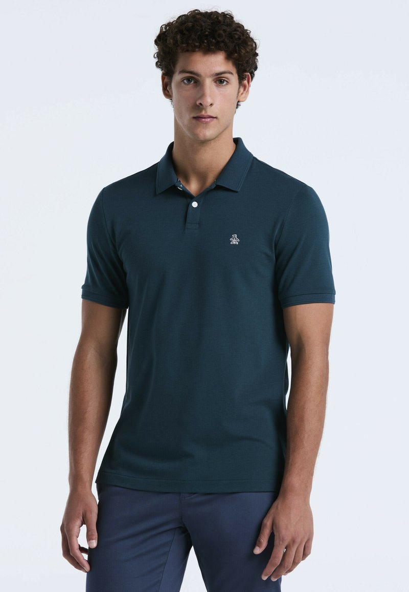 Junger Mann mit lockigem Haar, der ein dunkelblaues Polohemd und eine marineblaue Hose trägt und vor einem schlichten hellen Hintergrund steht.