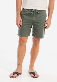 Groene katoenen short met een elastische tailleband en trekkoord, voorzien van zijzakken. Paar donkere teenslippers met een gevoerde zool.