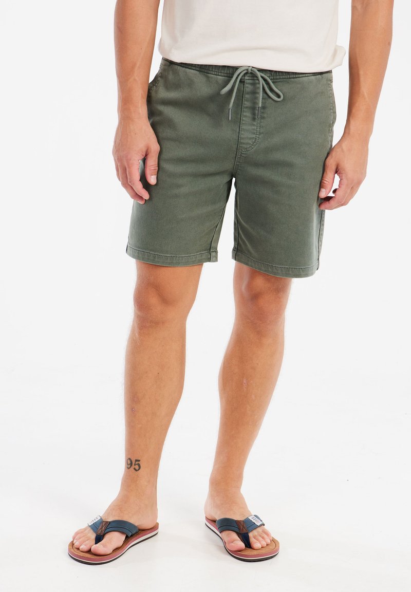 Groene katoenen short met een elastische tailleband en trekkoord, voorzien van zijzakken. Paar donkere teenslippers met een gevoerde zool.