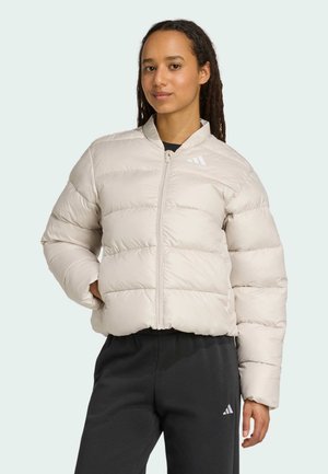 Jeune femme avec des dreadlocks portant une veste matelassée beige clair et un pantalon noir, posant devant un fond pâle.