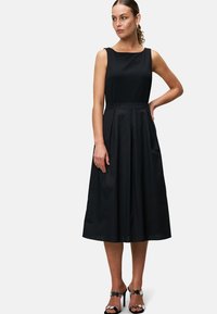 Black sleeveless dress with a fitted bodice and pleated, knee-length skirt. Smooth fabric with a subtle sheen. Paired with black heeled sandals.