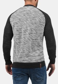 Hombre vestido con un suéter de mangas largas gris y negro y jeans azules, de pie de espaldas a la cámara sobre un fondo blanco.