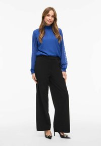 Blusa blu a chevron con collo alto, abbinata a pantaloni neri a gamba larga e tacchi a punta neri con accenti dorati.