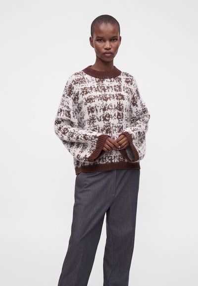 Personne portant un pull ample blanc et marron à motif avec poignets et col côtelés, associé à un pantalon gris foncé à jambes larges sur un fond uni.