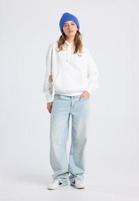 Woman wearing a blue knit hat, white hoodie with heart logo, light blue wide-leg jeans, and white sneakers standing against a plain background.