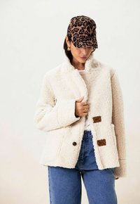 Veste en polaire beige, douce et fourrée, avec un col, une fermeture à boutons et deux poches latérales. Portée avec une casquette à motif léopard.