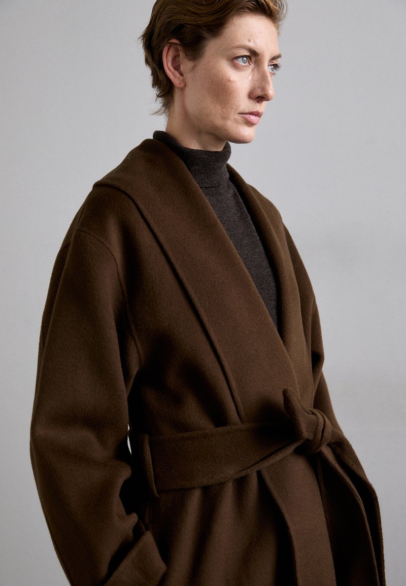 Cappotto avvolgente marrone in lana con colletto e vita regolabile. Caratterizzato da maniche lunghe e una texture morbida. Indossato sopra un dolcevita scuro.