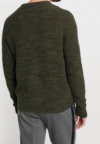 Homme portant un pull en maille texturée vert foncé et un pantalon de survêtement gris avec une bande noire, vu de dos sur un fond uni.