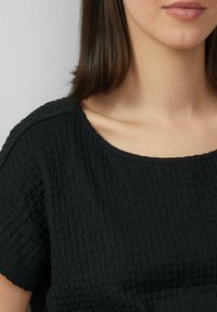 Top negro con una tela texturizada y arrugada. Presenta un escote ancho y redondeado, así como mangas cortas. Textura suave, diseño simple.