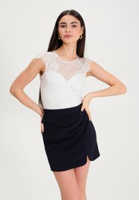 Body bianco in pizzo con maniche in rete trasparente, abbinato a una mini gonna drappeggiata blu navy con uno spacco laterale e un accento a fiocco davanti.
