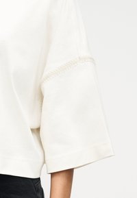 Κρεμ λευκό πουκάμισο με κοντά μανίκια και εμφανές ραφές στη μανσέτα, φορεμένο με σκούρο παντελόνι, σε απλό φόντο.