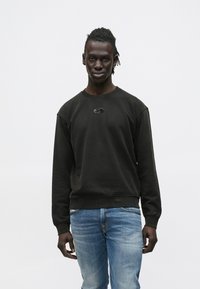 Μαύρο φούτερ με στρογγυλή λαιμόκοψη και ριμπ μανσέτες, με μικρό γκρι λογότυπο στο στήθος. Φορεμένο με μπλε τζιν.