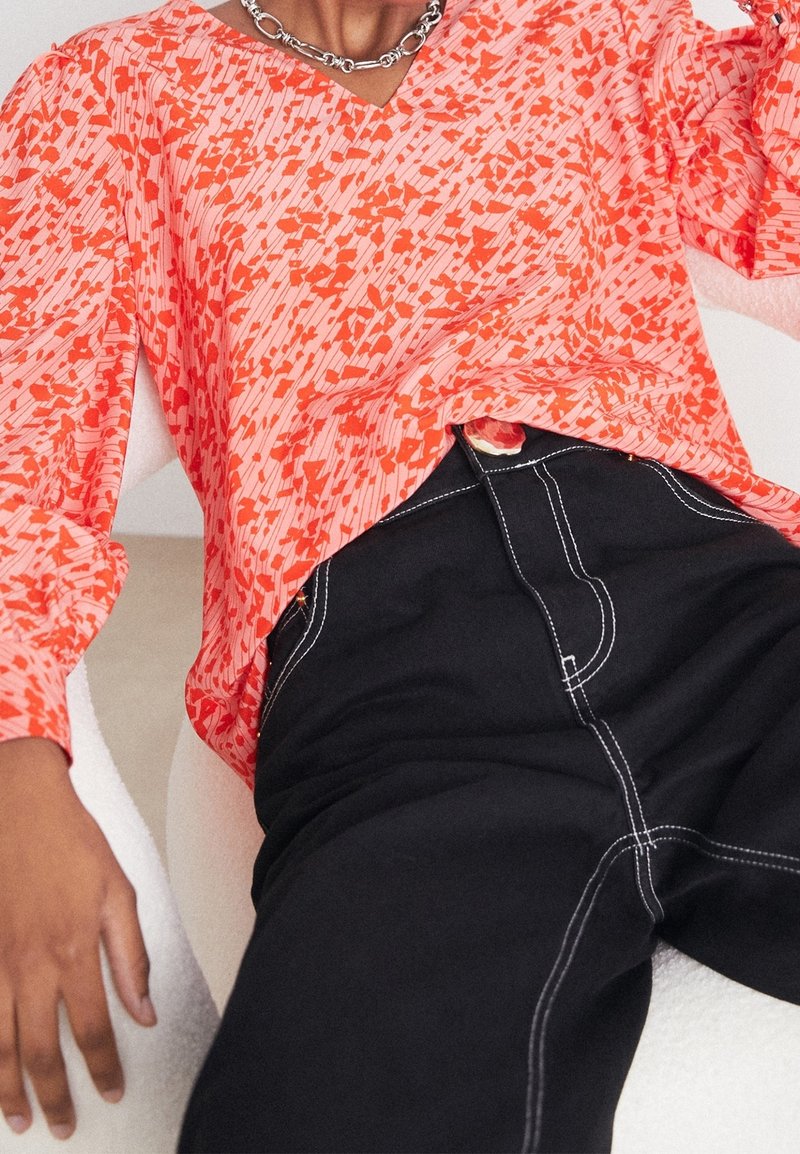 Bluse mit Korallenmuster, V-Ausschnitt, langen Ärmeln und lässiger Passform, kombiniert mit hochgeschnittenen schwarzen Hosen mit weißer Naht.