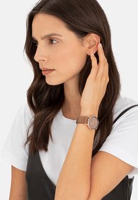Braune Lederuhr mit einem runden Gehäuse aus roségold und einfachem Zifferblatt. Modell trägt ein weißes Hemd und ein schwarzes Kleid, während sie sich die Haare zurückhält.