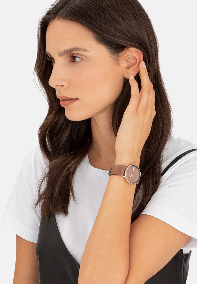 Braune Lederuhr mit einem runden Gehäuse aus roségold und einfachem Zifferblatt. Modell trägt ein weißes Hemd und ein schwarzes Kleid, während sie sich die Haare zurückhält.