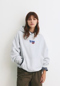 Jasnoszara bluza z kapturem z grafiką "Too Sweet" w niebieskim i czerwonym kolorze, z kangurzą kieszenią oraz ściągaczami na rękawach. Noszona na granatowej koszuli w kratkę.