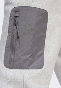 Grå idrottsbyxor med en strukturerad mörkgrå zipficka som har en präglad Adidas-logotyp och en mjuk tygfinish.