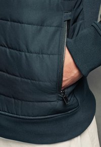 Veste matelassée bleu foncé avec une poche zippée, des poignets et un ourlet côtelés, présentant une texture lisse et un motif matelassé horizontal.