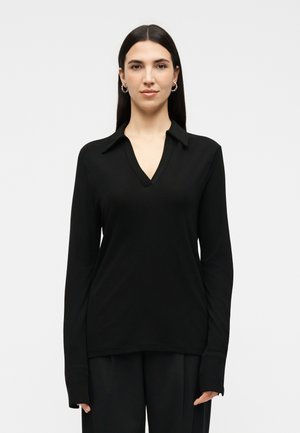 Γυναίκα με μακριά σκούρα μαλλιά, φορώντας χρυσούς κρίκους στα αυτιά και μαύρο μπλουζάκι με μακριά μανίκια και V- λαιμόκοψη, στέκεται μπροστά σε ένα απλό φωτεινό φόντο.